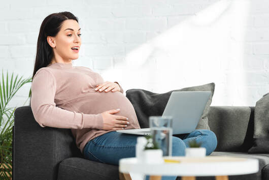 Happy pregnant woman sitting on the couch with her laptop during an online birth planning session with Elemental Beginnings. Adelaide Doula